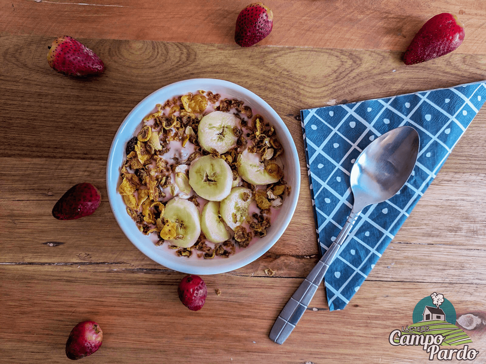 Pink yogurt bowl with granola and banana slices on a wooden table with strawberries.