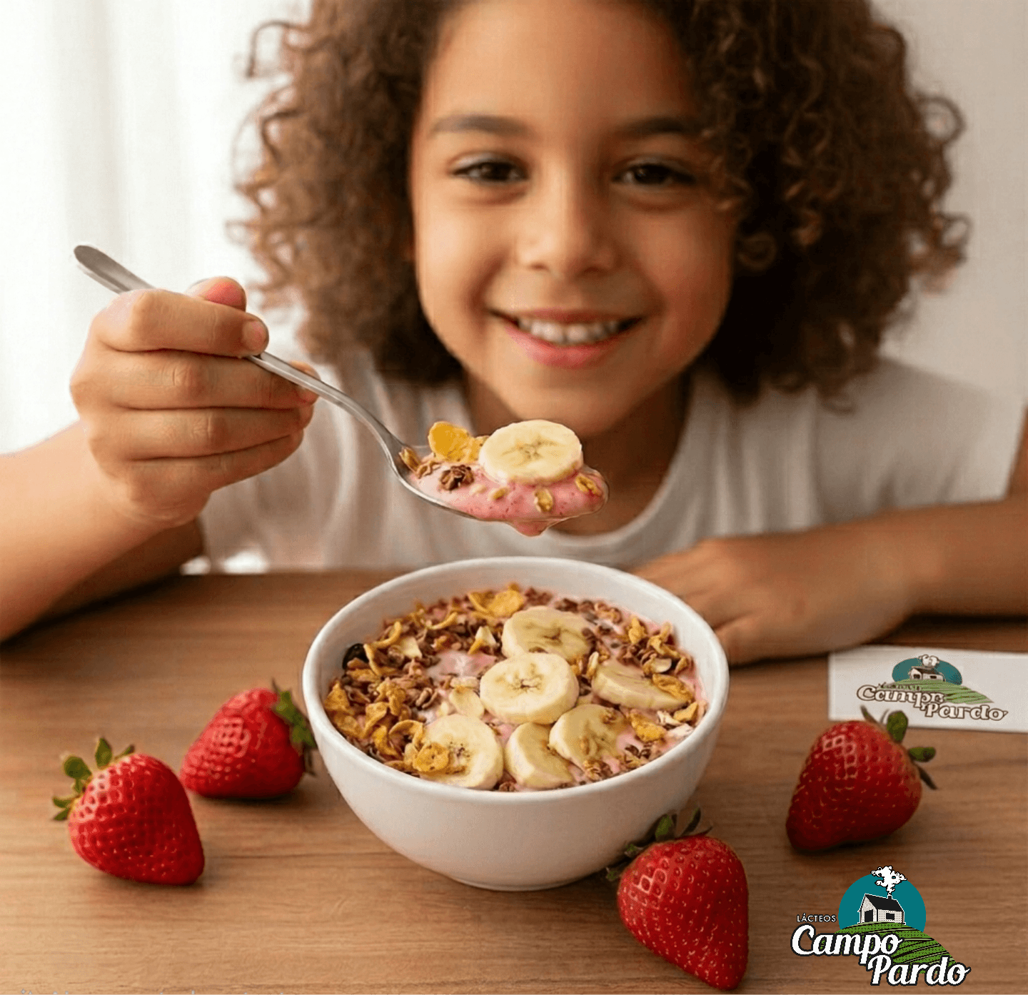 Smiling girl with curly hair eating pink yogurt topped with granola and banana slices.